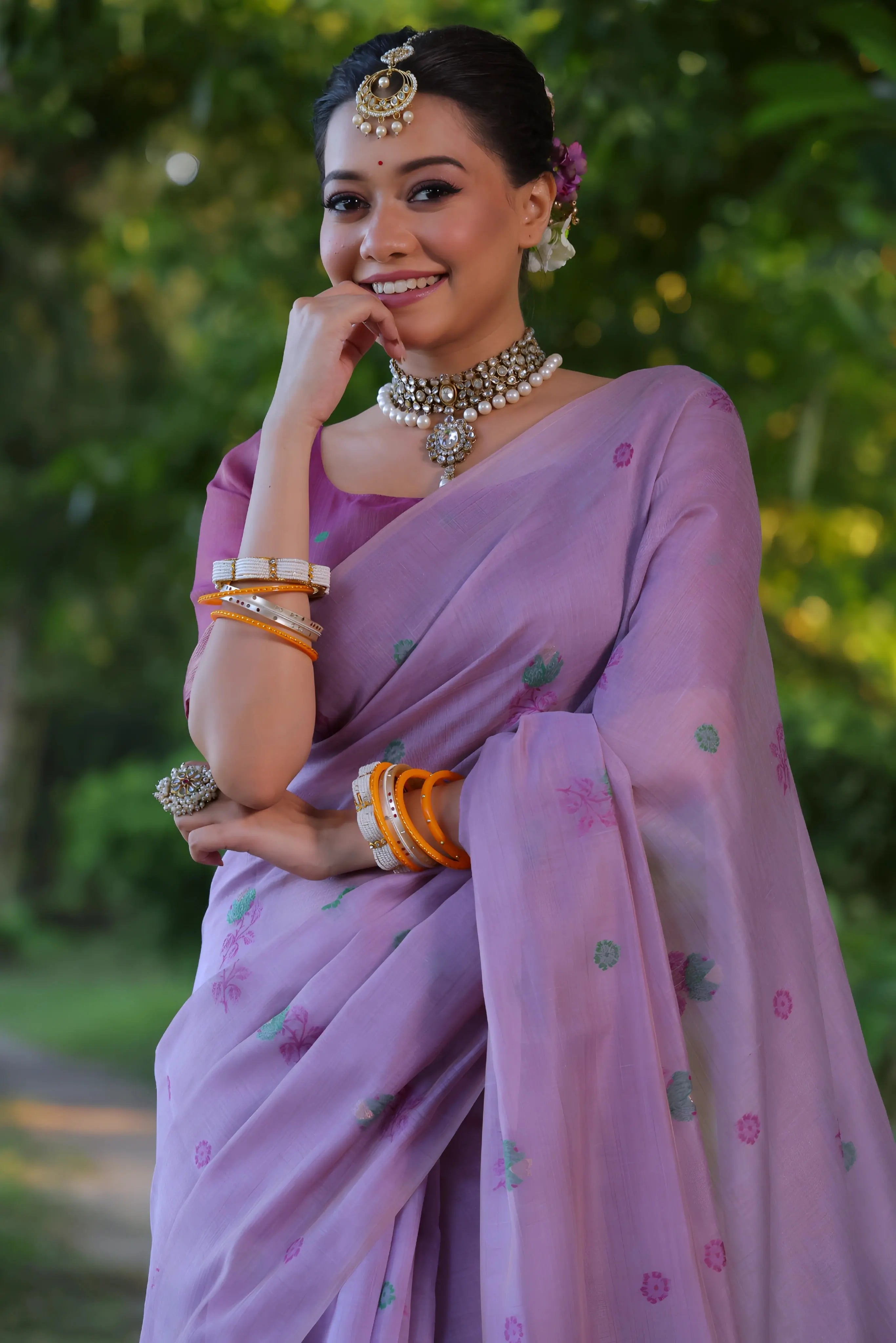A woman models an elegant light Lavender Soft Muga Cotton Saree with scattered floral Resham weaving. The pallu features an elaborate panel of multi-colored floral weaving/print in green and blue. She wears a contrasting deep maroon blouse, white stone/Kundan jewelry, and vibrant orange and yellow bangles, posing outdoors.